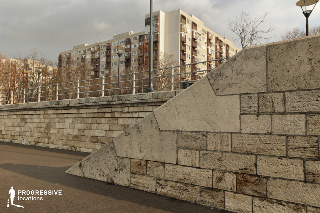 This image features a textured limestone embankment with a geometric, angular design, reinforced by metal railings along the top. The stonework exhibits natural variations in tone and texture, adding depth to the structure. In the background, a row of mid-century residential apartment buildings with repetitive window and balcony patterns contrasts with the classical stone construction in the foreground. The presence of streetlights and urban infrastructure further emphasizes the contrast between historical and modernist architecture. This location provides a dynamic setting for urban storytelling, architectural compositions, or social commentary on cityscapes.