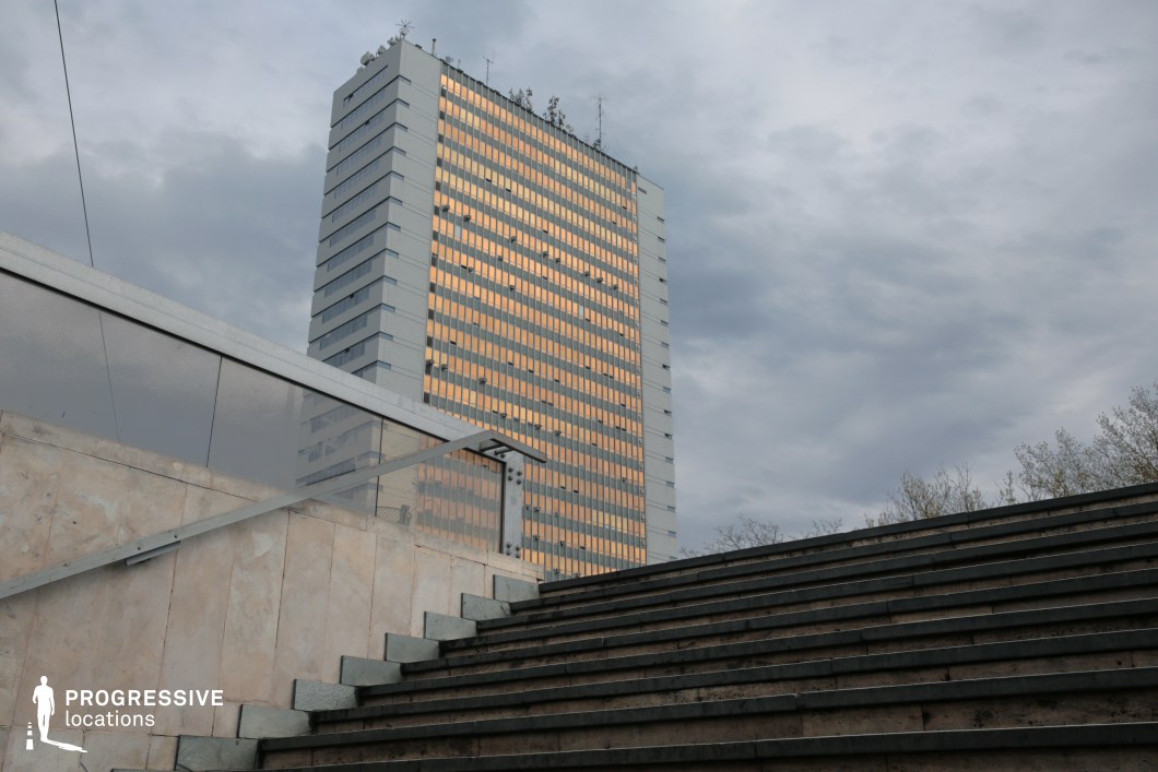 This image presents a contemporary urban scene, juxtaposing an angular stone staircase with a towering glass-clad high-rise in the background. The stairs are framed by a sleek metal and glass railing, enhancing the modernist aesthetic. The façade of the skyscraper features a grid-like pattern of reflective windows, casting a warm glow that contrasts with the cool tones of the stone and metal elements in the foreground. The shot’s composition emphasizes depth and scale, making it ideal for architectural, urban, or corporate-themed visual storytelling. The stark lines and geometric forms add a dynamic quality, perfect for cinematic or editorial use.