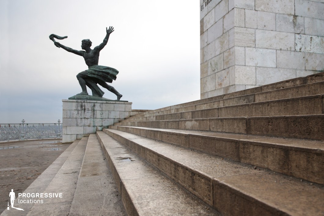 This image showcases a dramatic bronze statue in a dynamic pose, set against a grand stone monument. Positioned atop a raised platform, the statue depicts a figure with outstretched arms, exuding movement and power. The foreground features a sweeping limestone staircase, its worn surfaces adding a sense of history and permanence. The surrounding stonework is constructed from large, uniform blocks, reinforcing the monumentality of the space. The setting conveys themes of resilience, triumph, and historical significance, making it an ideal location for cinematic, historical, or symbolic visual storytelling.