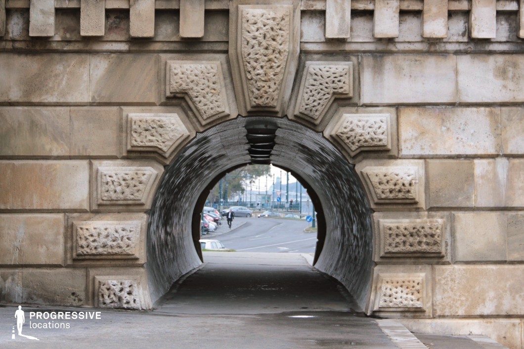 This image captures an intricately designed limestone tunnel built into a historic bridge or wall structure. The tunnel's arched entrance is framed by robust, textured stone blocks with geometric relief carvings, adding depth and character. The tunnel's interior has a darkened patina, contrasting with the bright exit revealing an urban street with parked cars and pedestrians. The perspective creates a natural framing effect, drawing the viewer's eye through the tunnel toward the modern cityscape beyond. This setting is ideal for cinematic urban sequences, architectural studies, or visual storytelling that juxtaposes historical craftsmanship with contemporary movement.