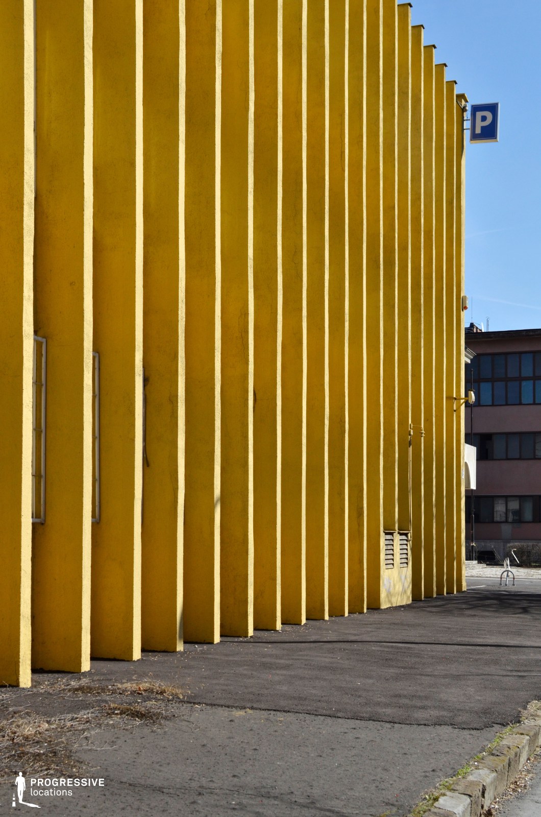 A bold architectural facade featuring a repetitive pattern of deep vertical grooves in a vibrant yellow hue. The structured design creates a strong geometric effect, with sharp contrasts between light and shadow. Small, narrow windows punctuate the surface, adding subtle details to the otherwise monolithic wall. The adjacent pavement is simple, leading toward a modern urban backdrop with mixed-use buildings. This location is ideal for conceptual photography, urban design studies, or as a visually striking backdrop for fashion and commercial productions.