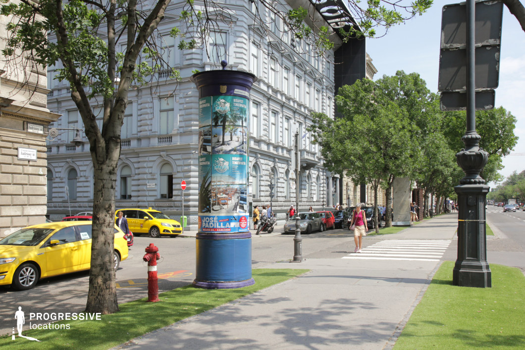 The image depicts a sophisticated urban boulevard characterized by a neoclassical building facade with ornate details and symmetrical windows. The dominant color palette consists of soft greys and whites, creating a refined and harmonious ambiance. A cylindrical advertisement column with vibrant posters contrasts against the understated tones, adding a dynamic visual element to the scene. The foreground features a tree-lined sidewalk with neatly maintained grass patches, which enhances the sense of order and cleanliness. A traditional street lamp, painted black and decorated with subtle ornamental designs, further emphasizes the classic aesthetic. The overall composition is open and spacious, ideal for capturing urban life or showcasing a mix of historical and modern elements in a film or photo shoot.