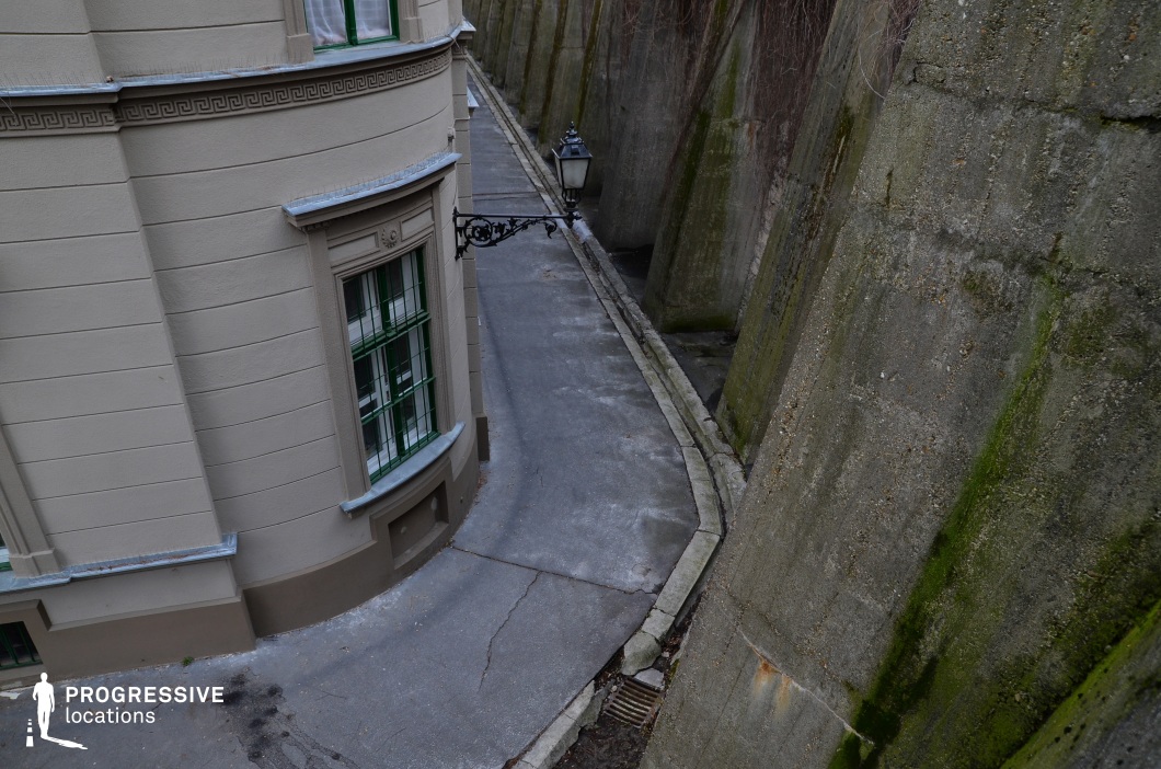 This scene presents an extraordinarily narrow walkway squeezed between two dramatically contrasting architectural elements. On the left, a finely detailed historic facade, featuring beige plaster, green-framed windows, and elegant ornamental molding, suggests a classical European urban setting. The adjacent towering wall on the right, constructed from weathered concrete with mossy streaks, strongly evokes the rugged structure of a fortress or city fortification, reinforcing the historical atmosphere. The alley's extreme narrowness amplifies the sense of confinement, with the pavement barely accommodating movement along its curved path. This juxtaposition of refined architecture and imposing defensive walls creates a unique, visually striking setting with a distinctly historical ambiance, perfect for scenes evoking tension or solitude.