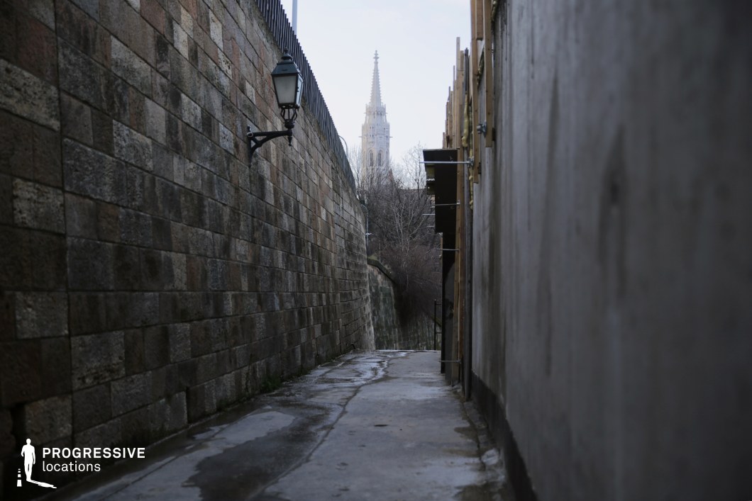 This image highlights a dark-toned, narrow alleyway that creates a sense of enclosure and inevitability, with no visible escape routes. On the left, a historic stone wall constructed from large, weathered blocks in earthy hues provides a timeless and rugged aesthetic. A black wrought-iron street lamp protrudes from this wall, enhancing the vintage atmosphere. The right side features a smoother, modern wall with exposed structural elements, adding an urban contrast to the scene.

The pathway is slightly uneven and damp, sloping gently downward toward a curve. Beyond the bend, the alley frames a majestic Gothic-style church spire in the distance, its intricate design standing as a striking focal point. The spire's elegant verticality contrasts with the alley's confined horizontal space, adding depth and character to the composition.

This dark-toned, narrow street, with its limited visibility and lack of escape routes, is an excellent choice for thriller or crime-themed locations, where a sense of tension and inevitability is essential.