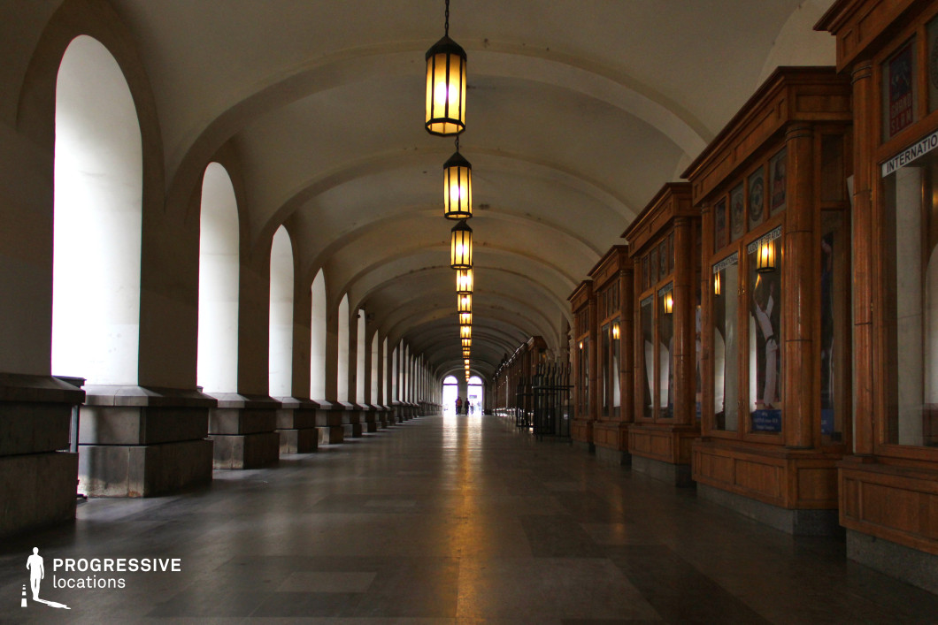 This image showcases a grand, arched arcade corridor, blending historic elegance with a sense of spacious symmetry. The left side features a series of tall, evenly spaced arches, supported by robust stone bases, allowing natural light to filter through the open spaces. The ceiling’s smooth curves are accentuated by a row of warm, hanging lanterns that illuminate the length of the corridor, creating a welcoming ambiance. On the right, ornate wooden display cases with glass panels and intricate detailing introduce a refined contrast to the stone elements. The polished flooring enhances the corridor’s formal aesthetic, while the linear perspective draws attention to the distant light at the corridor’s end, emphasizing depth and grandeur. This setting is ideal for highlighting architectural sophistication or creating a timeless atmosphere.