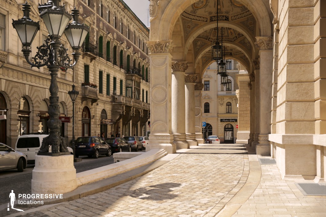 This image captures an elegant European street scene with a distinctly Parisian charm, featuring grand Neo-Renaissance architecture and intricately detailed colonnades. The ornate limestone arches with richly decorated ceilings evoke the sophistication of iconic Paris landmarks, while the classic black wrought-iron street lamps further enhance the Parisian aesthetic. The surrounding façades with arched windows, Juliet balconies, and green shutters add a quintessentially French touch. The cobblestone pavement and refined urban layout, reminiscent of Parisian boulevards, make this location ideal for film scenes set in 19th-century Paris or high-end fashion campaigns. Its timeless beauty, Parisian flair, and architectural richness provide a perfect backdrop for productions seeking elegance and historical authenticity.