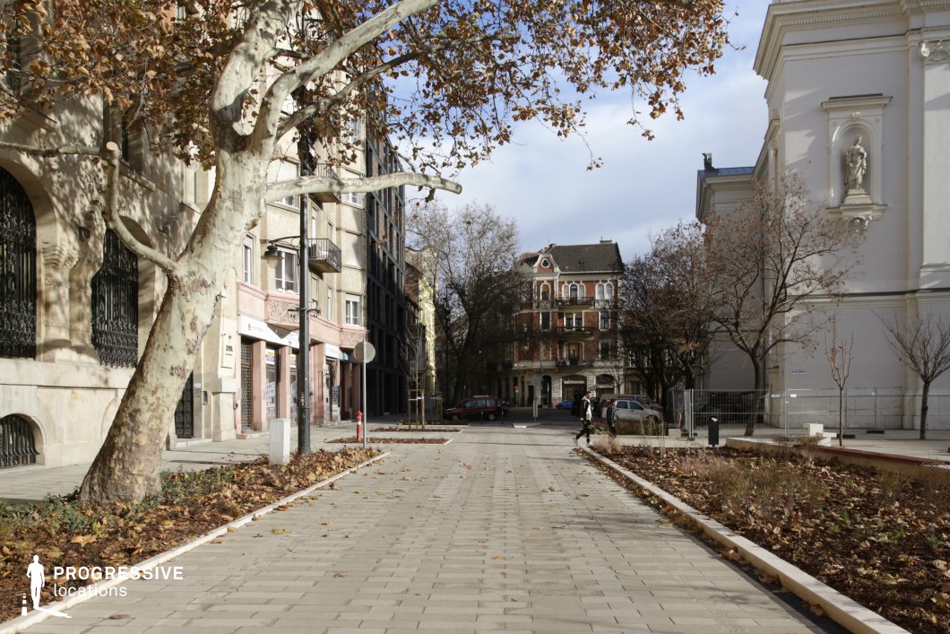 This image depicts a quiet European street with a pedestrian-friendly design, framed by historic architecture and seasonal autumn foliage. The centerpiece is a charming building with ornate balconies and colorful brickwork, complemented by surrounding structures featuring intricate wrought-iron details and stucco façades. The open square is lined with trees and newly landscaped greenery, creating an inviting, relaxed urban environment. The combination of historic and modern elements makes this location ideal for scenes requiring a picturesque, tranquil city setting, whether for period dramas, lifestyle shoots, or autumn-themed fashion campaigns. The warm natural lighting enhances the intimate, timeless atmosphere of the location.