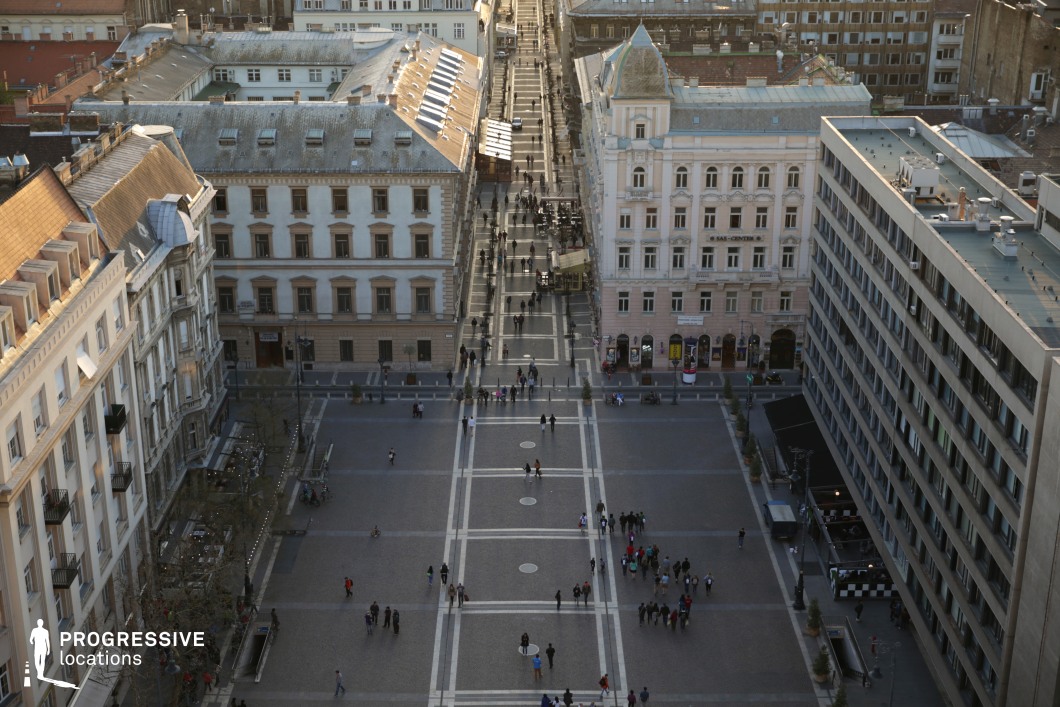 This image presents a bird's-eye view of a spacious pedestrian square, surrounded by a blend of classical and modern architecture. On the left, historic buildings feature ornate facades, pitched roofs, and wrought iron balconies, showcasing intricate detailing. On the right, a modernist building contrasts with its clean, minimalist lines and uniform appearance. The square itself is paved with a geometric pattern of light and dark stone, creating a sense of symmetry and order. The perspective draws attention to the central axis and the street extending beyond the square, lined with small cafes and trees. The combination of architectural styles and the open, well-defined space makes this location highly suitable for urban scenes in film, offering a dynamic yet elegant setting.