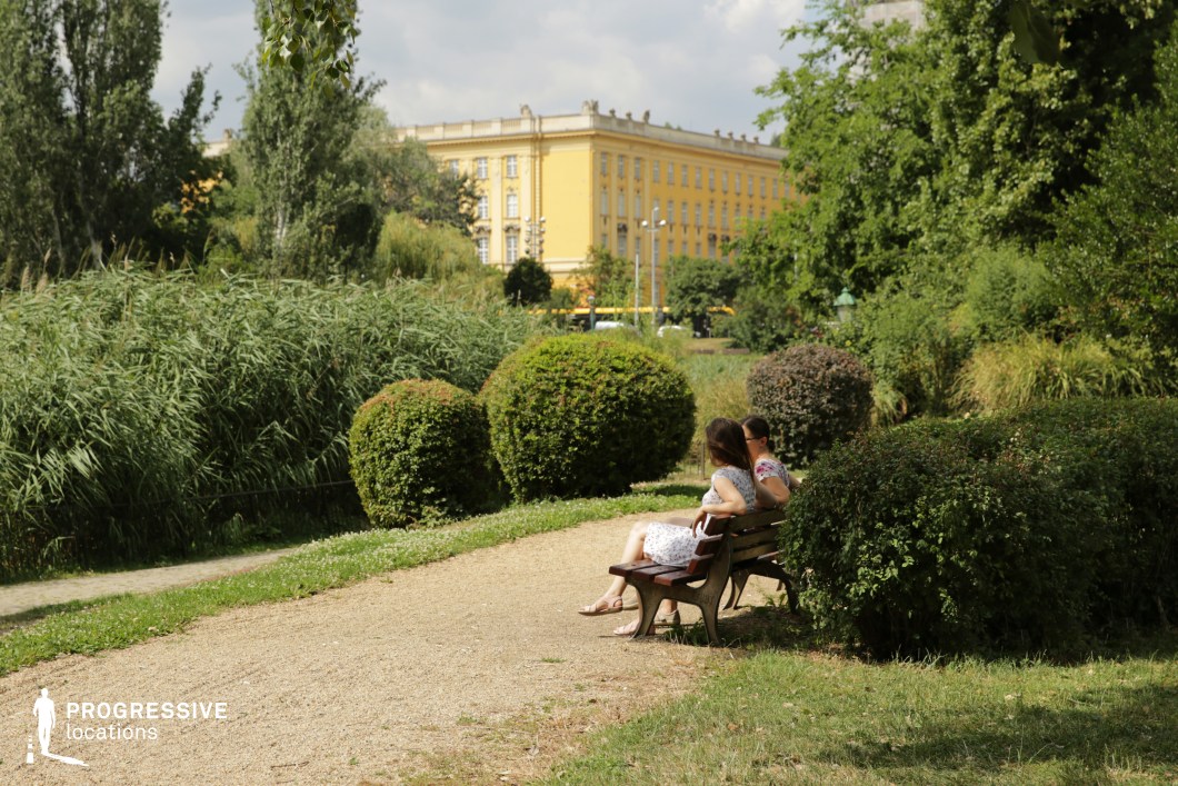 This image captures a tranquil park scene with a sandy pathway flanked by manicured greenery and lush trees, creating an intimate natural setting. In the background, a bold yellow neoclassical building adds a vibrant urban contrast to the organic tones of the park. The composition highlights the interplay between the structured, historical architecture and the soft, irregular forms of the bushes and trees. The park's inviting benches and open space offer a calm atmosphere suitable for reflective or romantic scenes. The soft, diffused light enhances the warm and peaceful ambiance, making this location ideal for filming serene, contemplative moments in an urban context.
