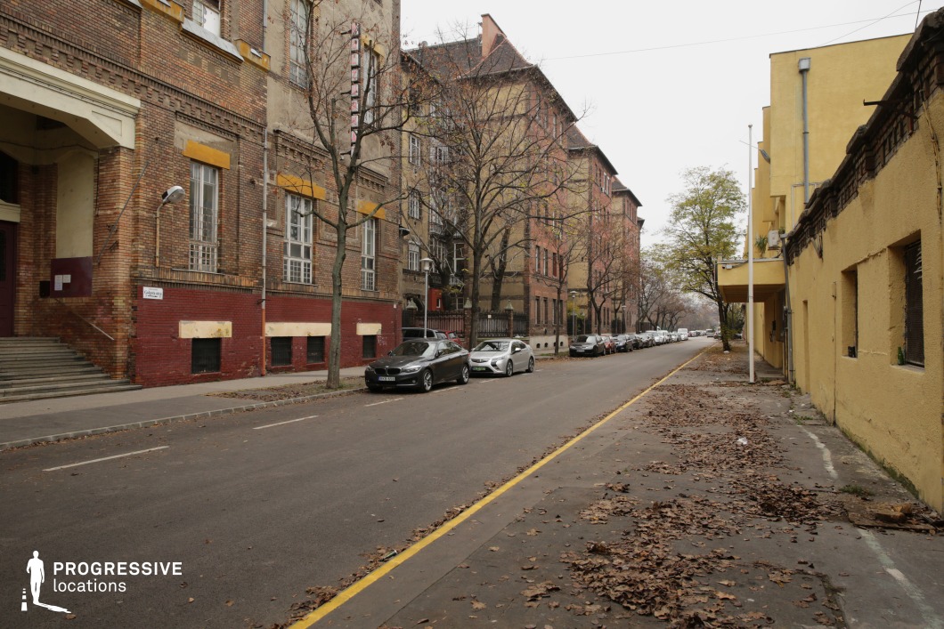 This image showcases a modest urban street bordered by buildings with warm brick facades. The structures feature a combination of red and brown brickwork, accented with yellow details around the windows and doors, creating a rustic yet vibrant character. The street is framed by rows of trees, enhancing the structured symmetry of the environment. On the right, a building with smooth yellow plaster provides a stark contrast to the textured brickwork of the left-side structures. The balanced blend of classic materials and vivid tones gives the scene a timeless, understated charm, suitable for capturing an everyday yet historically rooted urban setting.