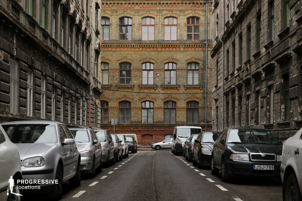 This location captures a narrow, enclosed urban street flanked by mid-rise historic buildings on both sides. The buildings exhibit richly textured stone and brick facades, marked by tall, evenly spaced windows and decorative cornices, reflecting a 19th-century architectural style. The street culminates in a three-story structure of warm-toned yellow brick, accented with red brick and arched window frames, which forms a visually dominant backdrop and architectural focal point. The visual palette is primarily composed of earthy tones—browns, greys, and muted yellows—creating a coherent and atmospheric setting. The use of aged brick, stone, and wooden window frames adds to the location’s historic and somewhat austere urban character. Vertical and linear geometric elements, such as window alignments and the road’s central perspective, define the visual rhythm and structure. Due to its symmetrical layout, strong central vanishing point, and enclosed proportions, this location is highly suitable for controlled filming environments, especially for urban scenes requiring a period-appropriate European backdrop with a dense, immersive city feel.