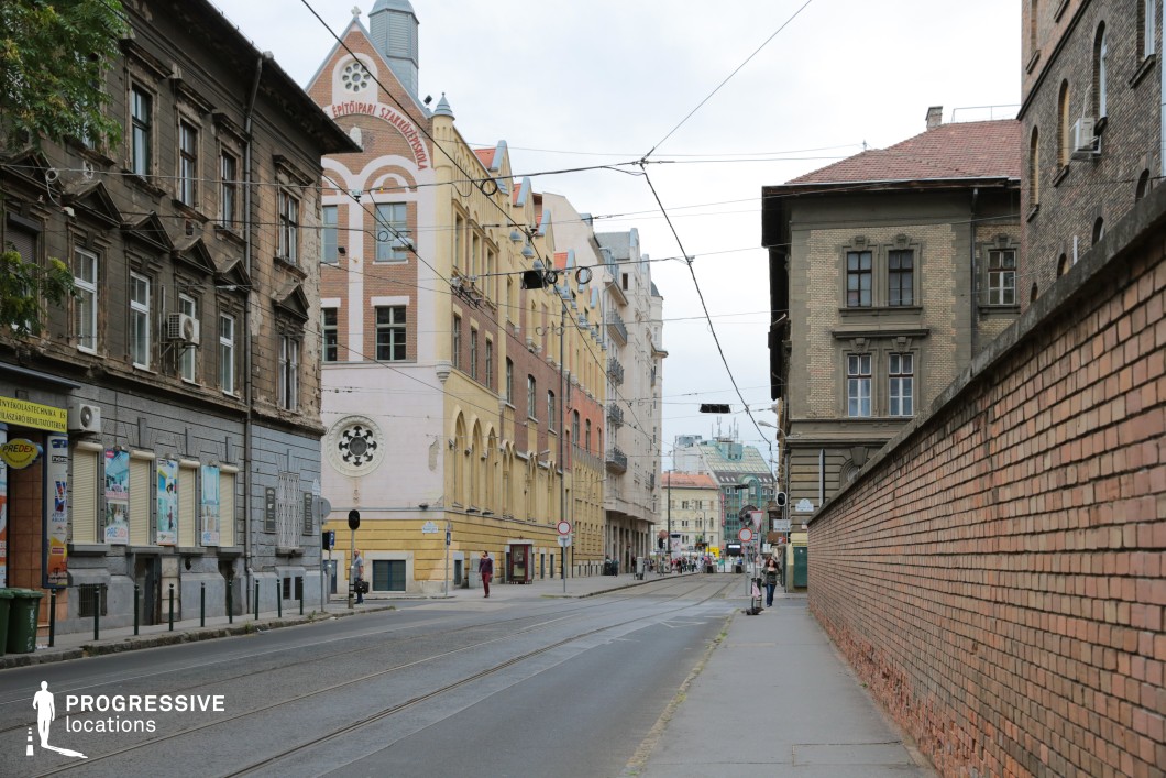 This image captures a historic street lined with diverse architectural styles, blending rustic charm with vibrant urban life. On the right, a long brick wall adds a sense of continuity and texture, contrasting with the ornate and weathered facades of the buildings on the left. A standout feature is the red-brick and cream structure in the center, adorned with pointed gables and a decorative circular window, evoking early 20th-century craftsmanship. Overhead, tram cables crisscross the sky, hinting at the functionality of the street as a transit route. The scene leads to a more modern cityscape in the distance, creating a layered composition of old and new. This location offers a versatile setting for films requiring a mix of historic character and urban realism.
