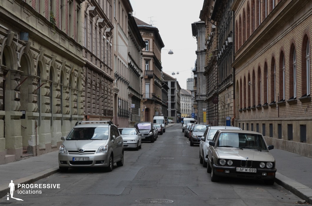 This image depicts a narrow urban street lined with historic buildings on both sides, characterized by intricate architectural details. On the left, decorative stone arches and textured facades showcase late 19th-century craftsmanship, while the right side features brick structures with evenly spaced arched windows, adding an industrial elegance to the scene. The composition emphasizes the street's linear perspective, leading the eye toward more distant buildings and creating a sense of depth. The muted tones and uniformity of the facades give the location a cohesive and timeless atmosphere, making it suitable for films or photography that require an intimate urban setting with European architectural charm.