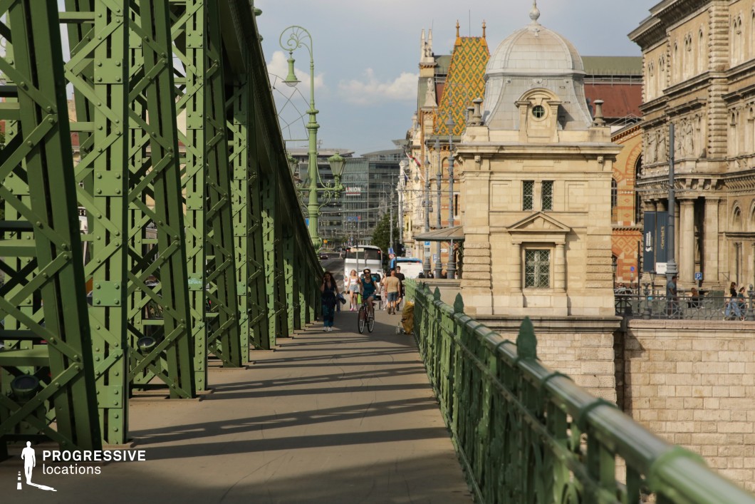 This image depicts a vibrant urban scene on a historic 19th-century bridge, showcasing the eclectic and Art Nouveau styles. The striking green ironwork of the bridge, adorned with ornate details and decorative lamp posts, reflects the aesthetic and technological advancements of the late 19th and early 20th centuries. The background features elegant buildings with colorful tiled roofs and intricate stone façades, embodying historicist elements typical of the era, blending Neo-Renaissance and Neo-Gothic influences. The lively pedestrian activity, with cyclists and walkers, adds a dynamic, modern contrast to the timeless architectural setting. This location is ideal for productions seeking 19th-century grandeur, industrial elegance, and bustling city life, as well as for capturing romantic scenes against a picturesque and historic backdrop.