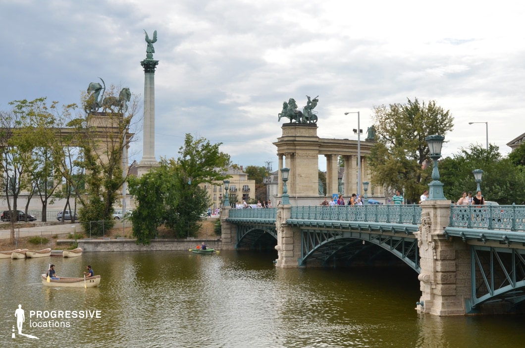 This image features a picturesque scene of a decorative bridge spanning a serene lake, framed by neoclassical architectural elements and monumental statues. The bridge, with its intricately designed wrought iron railings and stone supports, exudes elegance and historical charm. On the left, towering statues of mythological figures atop stone arches emphasize grandeur and artistic craftsmanship. In the background, a tall column crowned with a majestic angel statue adds verticality and a sense of monumental scale. The surrounding greenery and water provide a natural contrast to the ornate architectural details. This location is ideal for cinematic scenes that require a blend of historical grandeur and tranquil urban beauty.