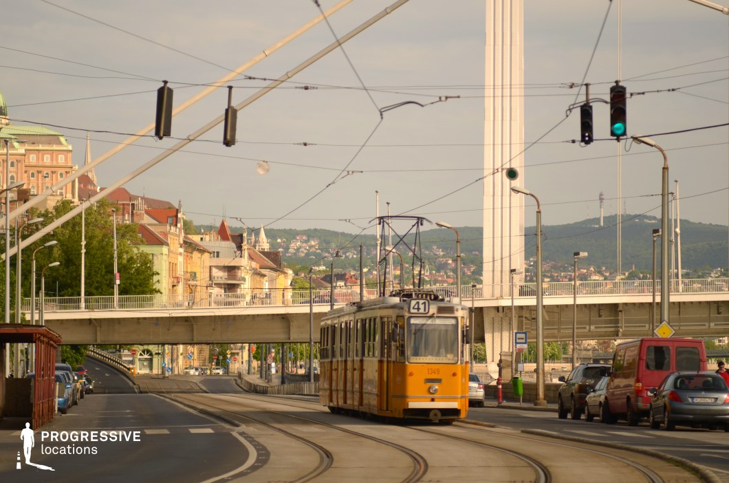 This image showcases a traditional yellow tram, a hallmark of historic European public transport, as it glides along its tracks beneath a modern bridge. The tram's classic design, featuring its distinct rounded edges and vintage character, contrasts with the more contemporary urban elements, such as the sleek bridge structure and overhead cables. In the background, the hillside is adorned with historic architecture, including castle-like structures, adding a sense of heritage and depth to the composition. The soft, golden light enhances the nostalgic charm of the scene, making this location ideal for productions seeking a blend of timeless urban transport and rich cultural backdrops.