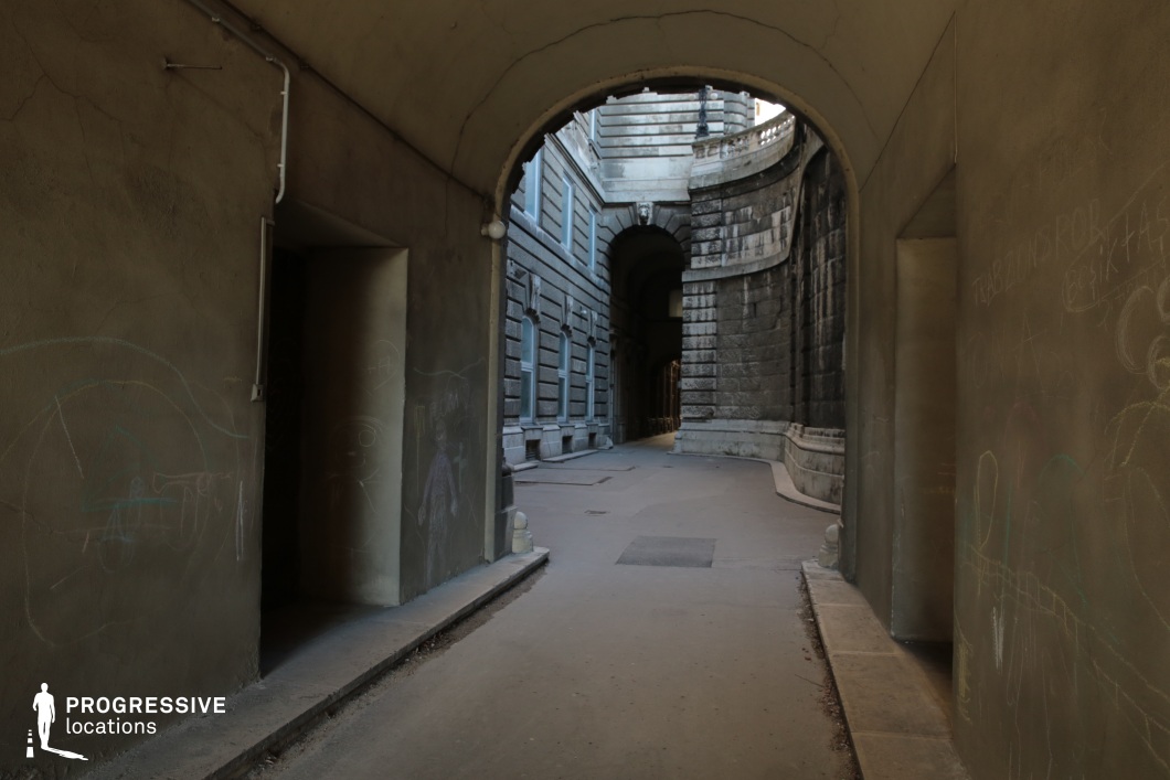 This atmospheric image captures a dimly lit arched passageway leading into a narrow alley flanked by massive, weathered stone walls. The foreground features plain, aged plaster walls marked with faint graffiti, contrasting the detailed and robust stone architecture visible in the distance. The curved pathway leads the eye towards the grand arched entrance further ahead, framed by classical stone detailing and ornate balustrades. The subdued lighting and confined perspective evoke a sense of mystery and seclusion, making the scene ideal for conveying historical depth or a dramatic, cinematic ambiance.
