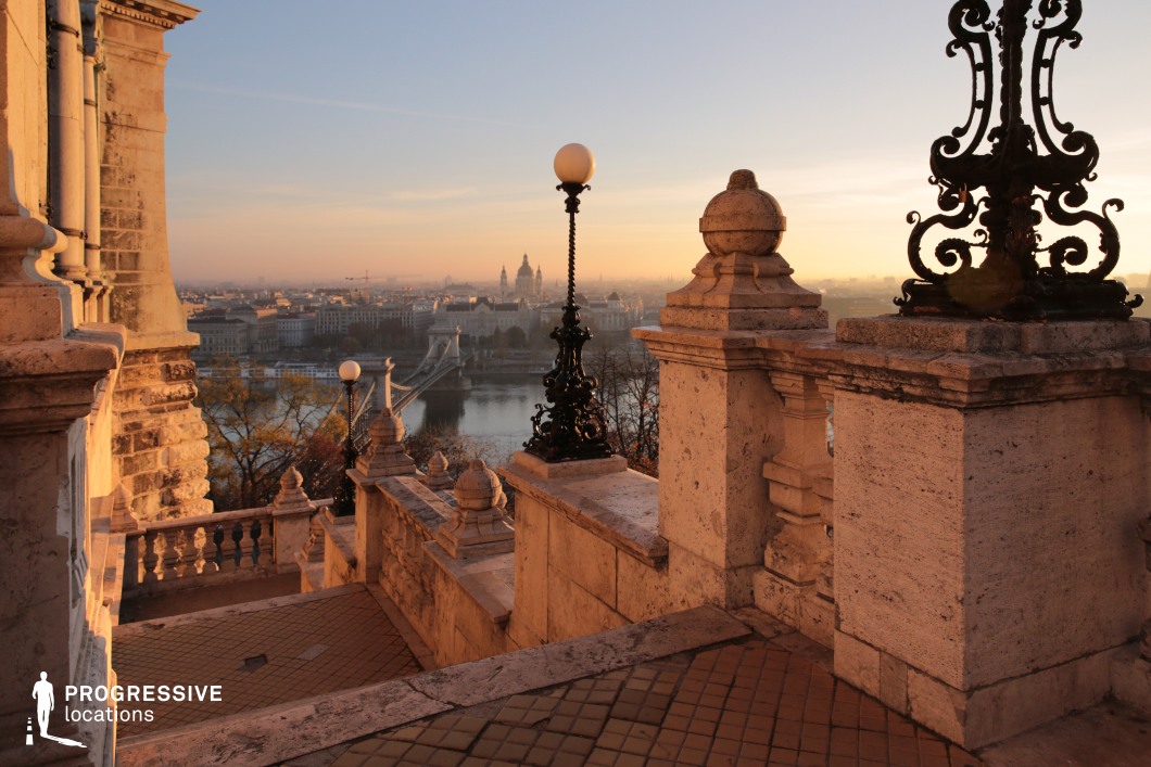 This captivating image showcases an ornate staircase descending from the heights of Buda Castle, offering a stunning panorama of Budapest. The balustrades, crafted from weathered stone, are adorned with intricate detailing, including sculpted finials and robust stone pillars. Elegant wrought-iron street lamps with globe lights add a touch of old-world sophistication, contrasting beautifully against the soft tones of the stone. Beyond the stairs, the iconic Chain Bridge spans the Danube River, leading the eye toward the distant cityscape, where the dome of St. Stephen’s Basilica rises prominently. The warm, golden hues of the setting sun enhance the scene's romantic and historical charm, making it ideal for dramatic and contemplative compositions.