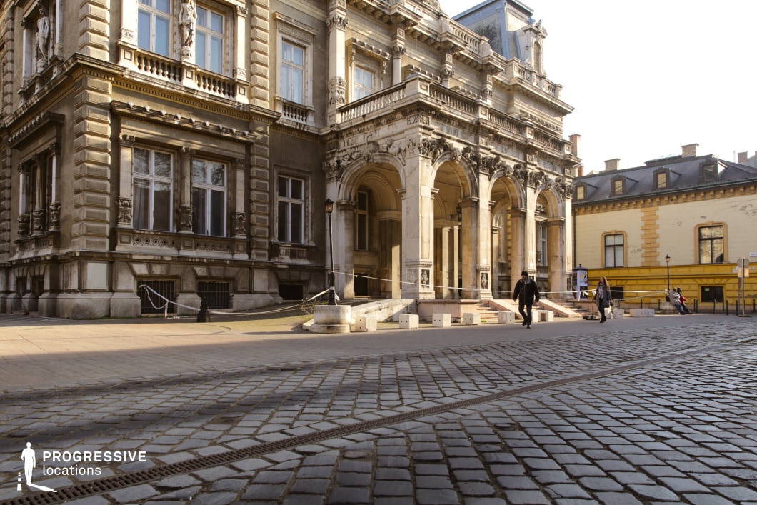 This image highlights an imposing classical building with ornate architectural details, including grand arches, intricate columns, and sculptural embellishments. The facade features a warm beige and stone-toned color palette, lending it a stately and timeless appearance. The cobblestone street in the foreground provides a textured, historic charm that complements the building's elegant design. To the right, a contrasting structure painted in vibrant yellow adds a subtle modern touch to the scene. The spacious open area and refined details make this location ideal for settings requiring a blend of historic grandeur and urban sophistication.