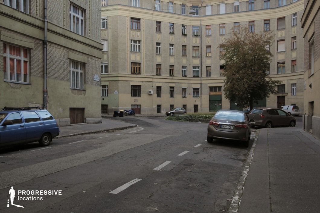 This image captures a medium-sized urban street lined with historic residential buildings. The facades are predominantly light brown and pale yellow, with subtle decorative details typical of mid-20th-century architecture. The slightly curved street layout adds depth and a sense of enclosure, while the large windows and simple geometric features highlight the buildings' functional design. A single tree near the center introduces a natural element, providing contrast to the urban tones. The weathered asphalt and muted colors of the buildings contribute to a calm, lived-in atmosphere, making this location suitable for productions requiring a historic yet understated residential setting.