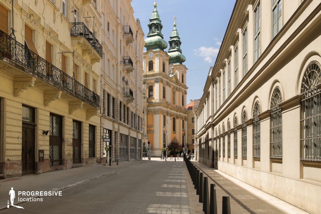 This location image presents a narrow, linear street flanked by historic urban architecture, culminating in a striking Baroque-style church with twin towers and green patinated domes. The symmetrical composition leads the viewer's gaze directly toward the ornate church façade, which features arched windows, columns, and elaborate stucco decorations in soft peach and ivory tones. The buildings on either side of the street showcase diverse yet harmonious neoclassical and Baroque-revival architectural elements. On the left, decorative wrought-iron balconies and detailed stone moldings add elegance and texture, while the right-hand façade is more austere with evenly spaced arched windows protected by traditional metal grilles. The dominant materials are stone and wrought iron, contributing to a historic and refined visual tone. The perspective from ground level emphasizes the verticality of the church and the enclosing nature of the street, creating a strong visual corridor. This location is ideal for productions seeking a European old-town ambiance, offering both architectural richness and a spatially intimate setting.