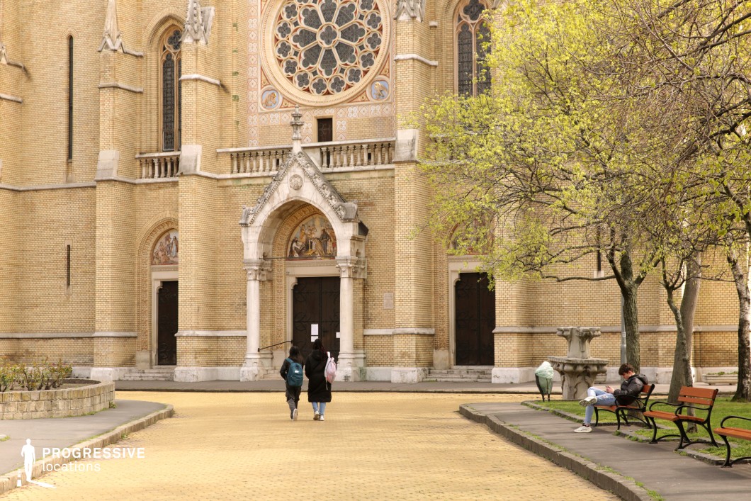 This image showcases a serene scene of a historic church with stunning yellow brickwork, intricate Gothic Revival details, and a prominent rose window as its centerpiece. The foreground features a tranquil courtyard with benches, trees just beginning to bloom, and a peaceful atmosphere perfect for reflective or intimate moments. The combination of the church's ornate façade and the inviting park-like setting makes this location ideal for period film scenes, romantic narratives, or lifestyle photography. Its timeless charm and architectural richness provide a striking backdrop for productions seeking elegance and authenticity.