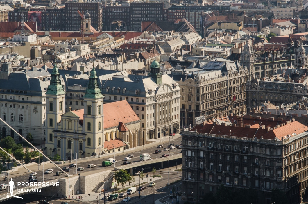 This panoramic view captures the architectural richness of Budapest's historic core, blending Baroque, Neo-Renaissance, and early 20th-century styles. Dominating the foreground is the Baroque-style Inner City Parish Church, with its pale yellow facade, twin towers topped with green patina domes, and red-tiled roof. Adjacent to it, the Neo-Renaissance buildings showcase ornate facades, symmetrical windows, and intricate stone carvings, reflecting the grandeur of 19th-century urban planning. The Elizabeth Bridge crosses the Danube River in the lower left, adding a modern element to the composition, while bustling traffic beneath it introduces a dynamic, urban character. Surrounding rooftops with varied designs and colors create a layered cityscape, emphasizing the city's architectural diversity and vibrancy. This scene is ideal for establishing a historic yet lively urban atmosphere in creative works.