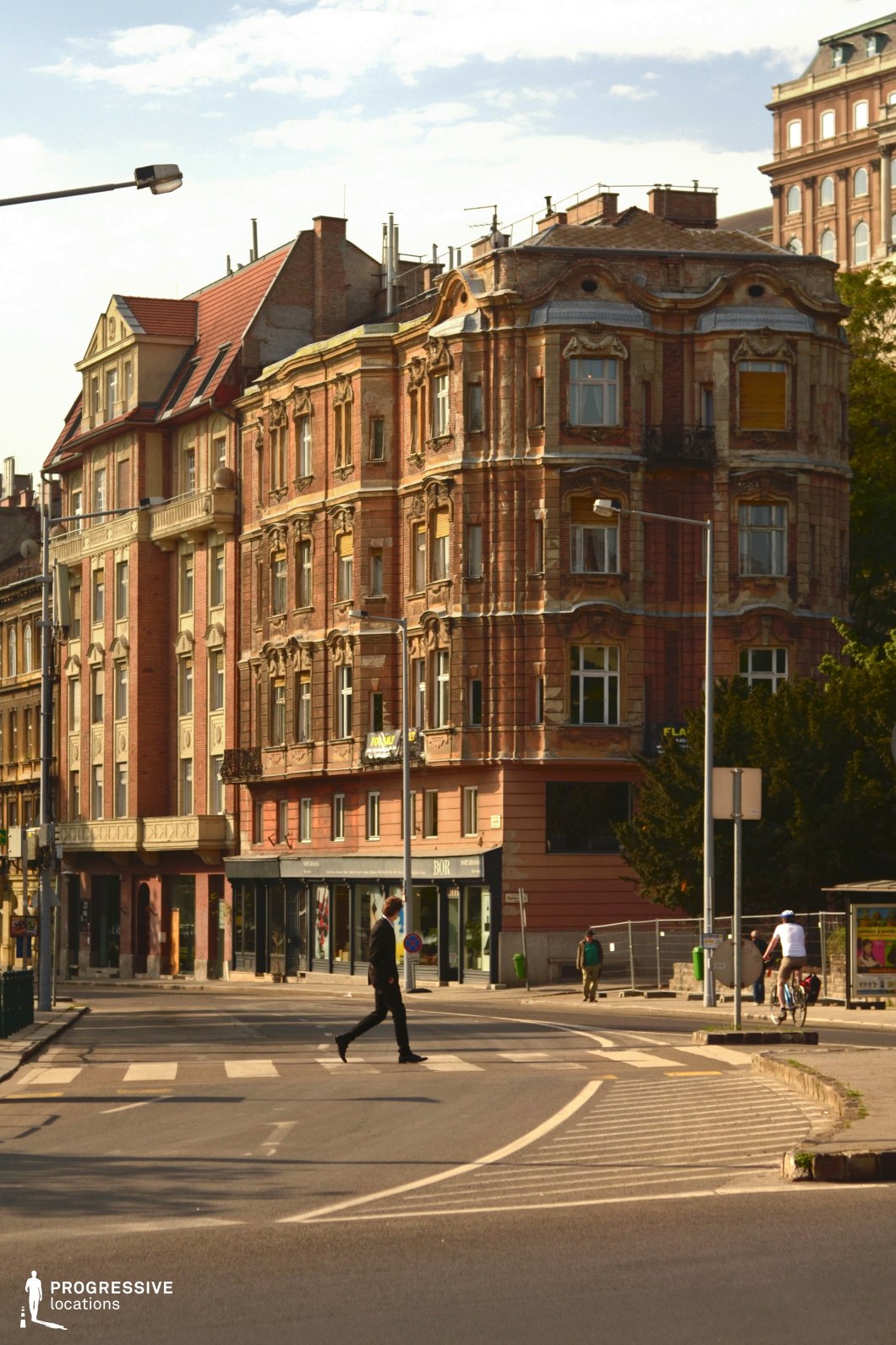 The focus of this image is a lively urban intersection, where a pedestrian crosswalk brings together a colorful and welcoming city atmosphere. Surrounding the street are richly detailed historic buildings, their warm reddish and beige tones adding to the scene's inviting aesthetic. The corner building, with its Neo-Baroque design and rounded facade, features ornate stucco work, arched windows, and a small wrought-iron balcony, giving the area a sense of architectural elegance. The combination of curved streets, urban greenery, and the dynamic presence of pedestrians and cyclists enhances the bustling, cosmopolitan vibe. This setting embodies a harmonious blend of historic charm and vibrant city life, ideal for capturing the energy and warmth of a modern metropolis.