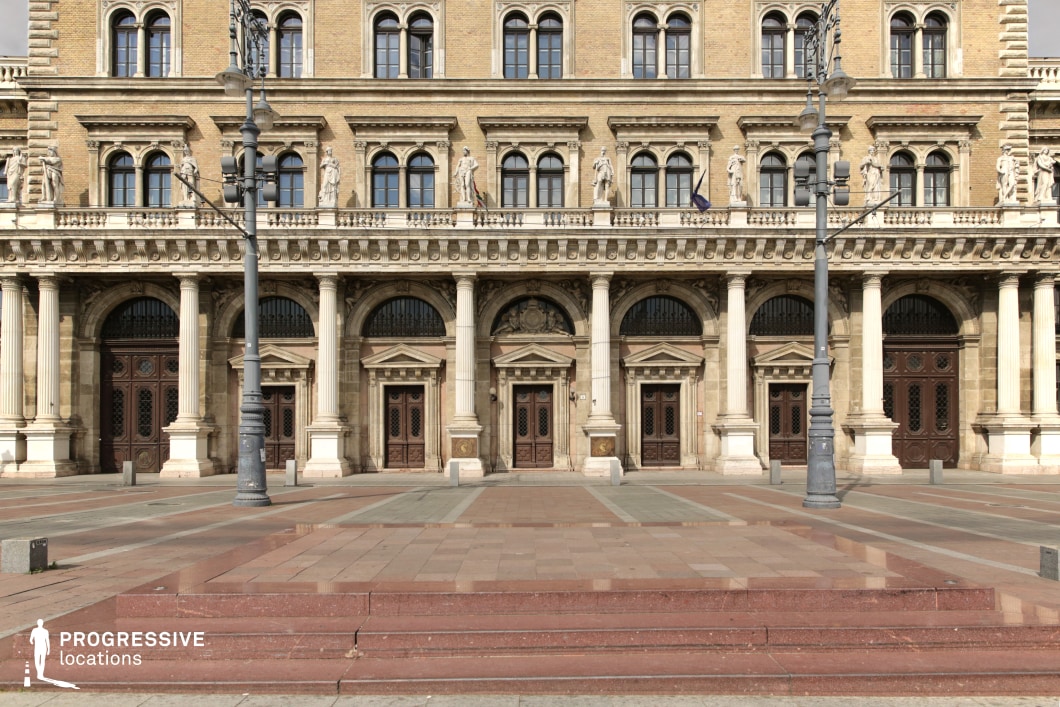This image showcases the grand façade of a Neo-Renaissance building from the 19th century, characterized by its symmetrical design and ornate architectural details. The structure features a series of imposing arched doorways framed by classical columns, with intricately carved statues and decorative elements adorning the upper level. The warm tones of the brick façade and the rich detailing of the doors and windows emphasize the building's elegance and historical significance. The open square in front of the building is expansive, offering ample space that enhances the monumental feel of the location. This wide plaza, with its carefully designed steps and clean lines, is ideal for cinematic shots of formal entrances, period dramas, romantic rendezvous, or grand public gatherings. The timeless grandeur and refined design evoke a distinctly European charm, reminiscent of iconic cultural and civic landmarks.