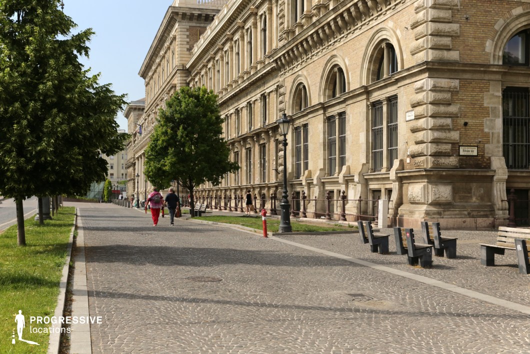 This image showcases a grand Neo-Renaissance building from the 19th century, characterized by its symmetrical design, arched windows, and richly detailed stonework. The building’s warm yellow brick façade, accented with beige and grey stone elements, creates a harmonious and elegant color palette that blends beautifully with the surrounding urban environment. The cobblestone street in the foreground adds earthy tones, while the lush green trees and classic black street lamps provide contrasting highlights that enhance the overall visual appeal. The wide pathway and open space, complete with public benches, offer a serene atmosphere, making it an inviting setting for capturing leisurely moments or grand entrances. This location is ideal for period dramas, romantic scenes, or historical narratives, offering not only architectural grandeur but also a sophisticated interplay of natural and urban tones.