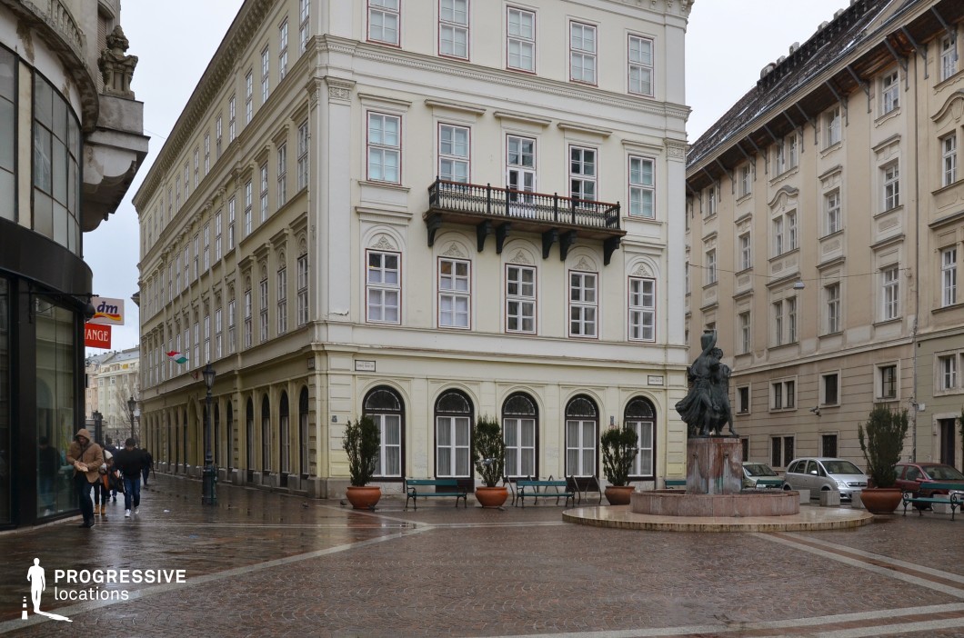 This image features a charming cobblestone square surrounded by elegant historic buildings. The central structure, painted in soft cream tones, is adorned with arched windows and a traditional balcony supported by intricate wooden brackets, evoking late 19th-century architectural styles. To the right, additional buildings complement the aesthetic with their harmonious design and subtle detailing. A bronze statue on a raised platform adds a focal point to the square, providing an artistic touch to the scene. The polished cobblestones, slightly damp, reflect light and enhance the square's inviting ambiance. This location is perfect for settings requiring a blend of historical charm and refined urban sophistication.