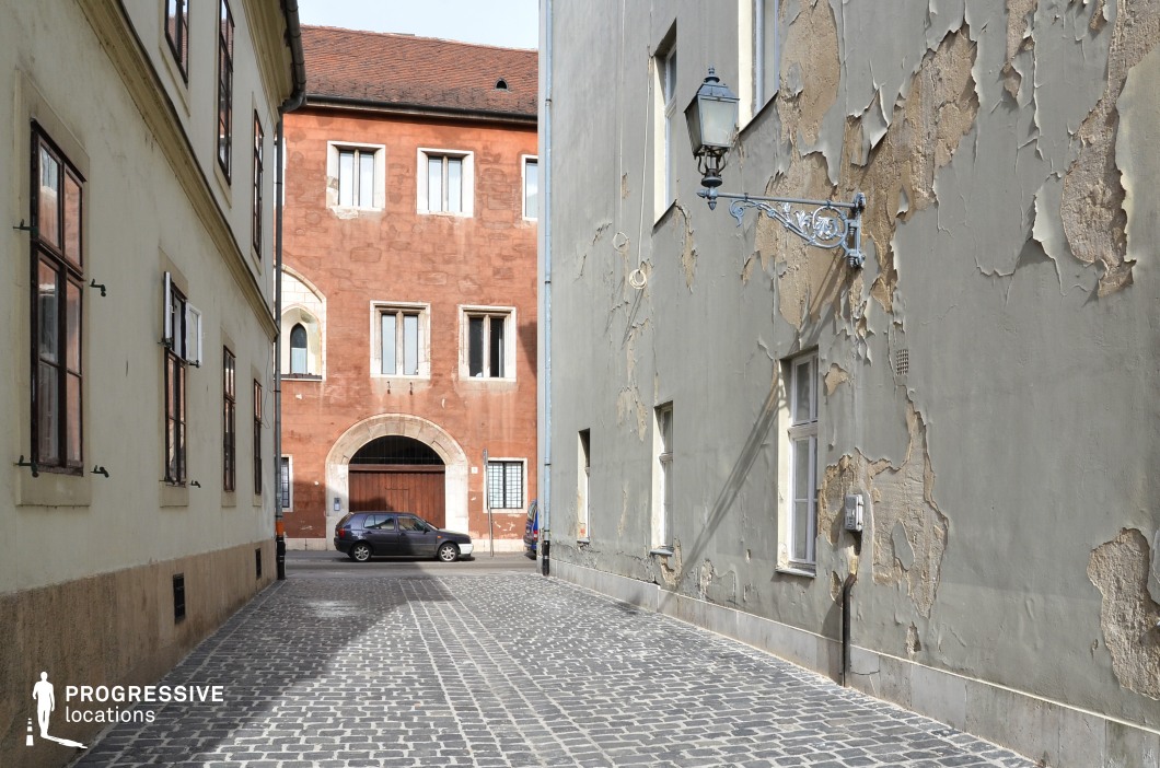 This image showcases a narrow cobblestone alley in a historic district, flanked by buildings with weathered facades that tell a story of age and character. On the left, a pale cream-colored building features wooden-framed windows with shutters, while on the right, a muted green wall with peeling plaster reveals layers of history. The alley opens up to a reddish-brown building with a large arched entrance and stone-framed windows, creating a striking focal point. A wrought iron lantern mounted on the wall adds a touch of classic charm. The textured surfaces and contrasting tones make this location ideal for productions requiring an atmospheric and authentic European old-town setting.