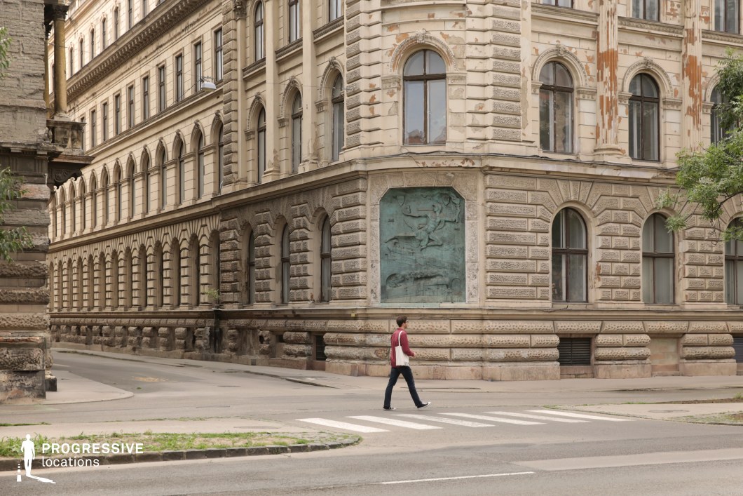 The image showcases a prominent corner building with a richly textured façade made of light beige stone. The structure features repetitive arched windows framed by detailed classical ornamentation, reflecting a historic European architectural style. A significant element is the large relief panel embedded on the corner wall, providing a focal point of artistic interest. The rusticated stonework at the ground level adds a robust and monumental feel, while subtle weathering and peeling elements on the upper sections enhance its aged character. The intersection of two streets creates dynamic visual lines, making this location suitable for dramatic, urban, or period-specific scenes.