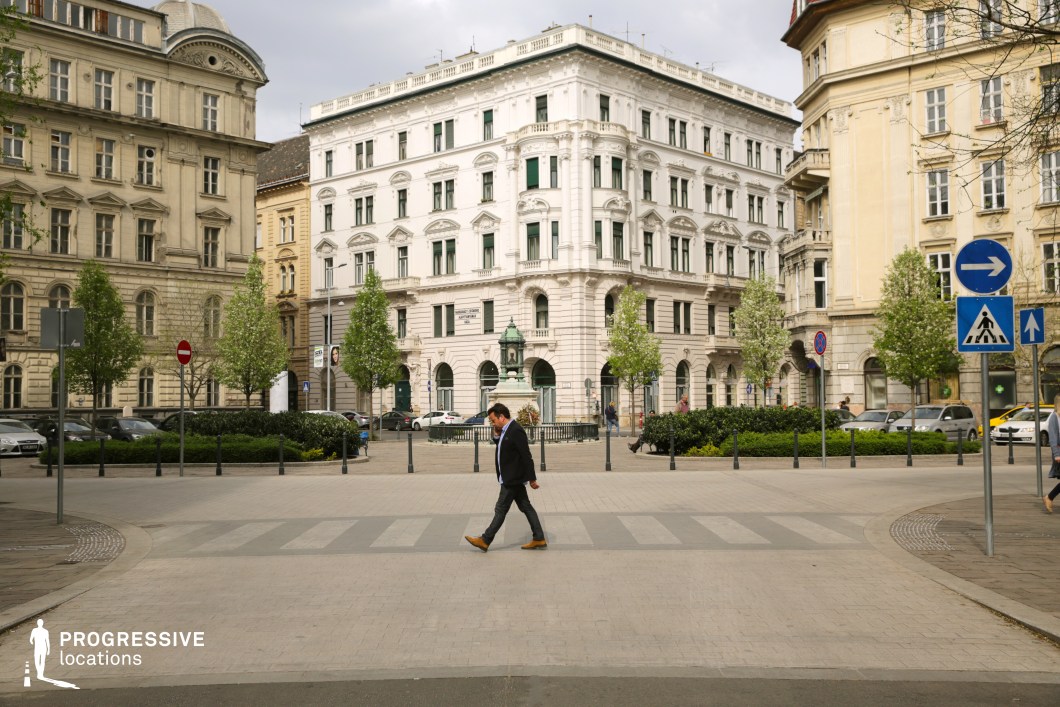 This image depicts a refined city square surrounded by grand historic buildings in a harmonious blend of neoclassical and late 19th-century architectural styles. The central white building stands out with its intricate ornamentation, arched windows, and symmetrical design, creating a focal point of elegance. The square itself is landscaped with lush greenery and young trees, bordered by well-maintained hedges that provide a soft contrast to the urban architecture. A decorative fountain or statue at the center adds a sense of sophistication to the space. The spacious pedestrian crosswalk in the foreground enhances accessibility and openness, making this location ideal for filming scenes that require a blend of urban vibrancy and historic charm.