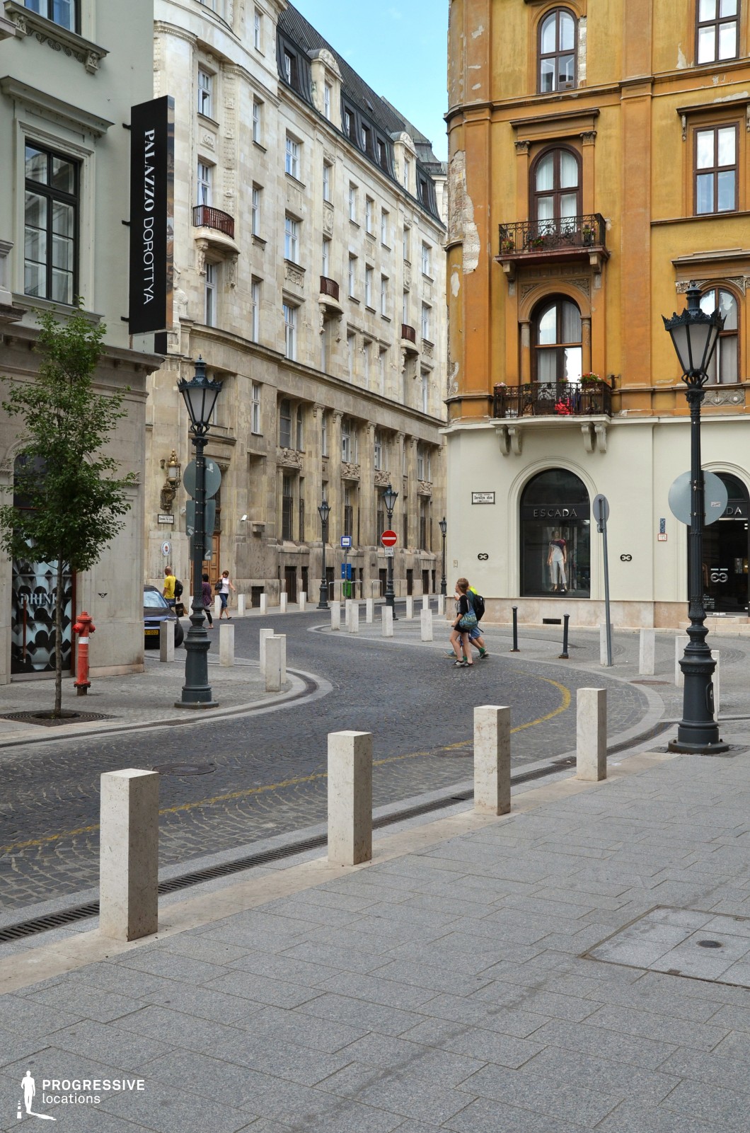 This image showcases a graceful urban street with a distinct curved design, lined with historic buildings that exude a classic European charm. On the right, a warm mustard-colored facade with intricate balconies and arched windows adds vibrancy and architectural depth. Opposite, a cream-toned building with elegant details and subtle accents provides balance to the composition. The cobblestone roadway enhances the historic ambiance, while modern white stone bollards and ornate black lampposts provide functional and aesthetic contrast. The intimate scale of the street and the well-preserved architecture create a refined, picturesque setting, ideal for highlighting urban sophistication and timeless design.