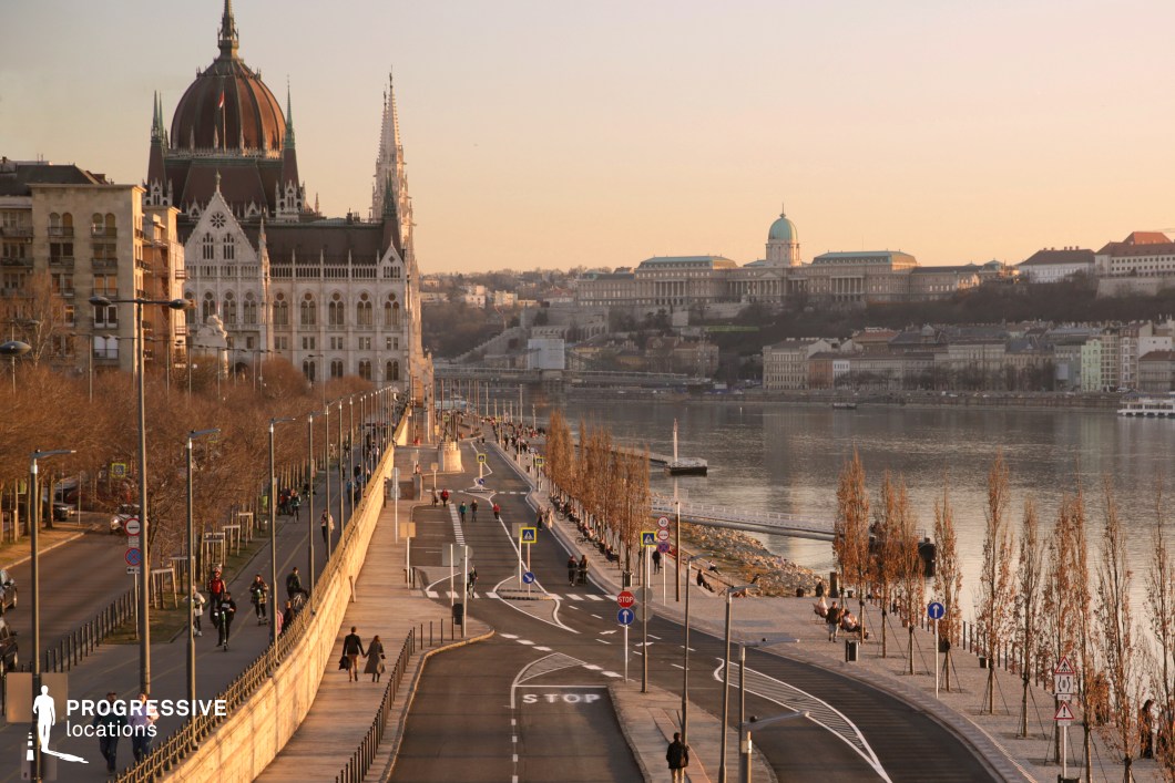 This image captures a dynamic riverside setting, focusing on the wide, intersecting pedestrian and cycling lanes that stretch along the promenade. The curving pathways provide a perfect foreground for filming scenes involving motion, whether it’s walking, cycling, or group interactions. The elevated perspective, likely taken from a bridge, adds a cinematic quality, emphasizing the spatial layout and the relationship between the roadways and their surroundings. In the background, the iconic Neo-Gothic Parliament building and the Buda Castle add grandeur and a sense of place, making the location visually compelling for productions. This combination of practical pathways and a historic urban backdrop offers a versatile setting for scenes requiring both movement and a strong sense of location.
