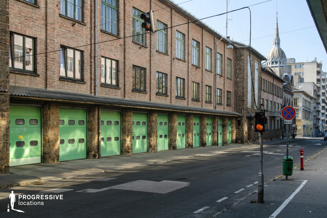 This image showcases a long brick fire station building with a utilitarian yet distinct early 20th-century industrial aesthetic. The facade is constructed from warm-toned brick and accented by tall, evenly spaced windows framed in dark metal. The row of large green garage doors at street level adds a vibrant touch, contrasting against the earthy tones of the brick and the stone columns between each gate. A corrugated roof overhang extends across the garage doors, emphasizing the functional design of the building. In the background, a pointed spire from a nearby historic structure introduces a striking architectural juxtaposition between industrial and ecclesiastical styles. The scene is completed by the quiet urban street with a traffic light and signage, capturing the harmony of practical infrastructure within a bustling cityscape. This setting is ideal for emphasizing functionality, history, and urban character.
