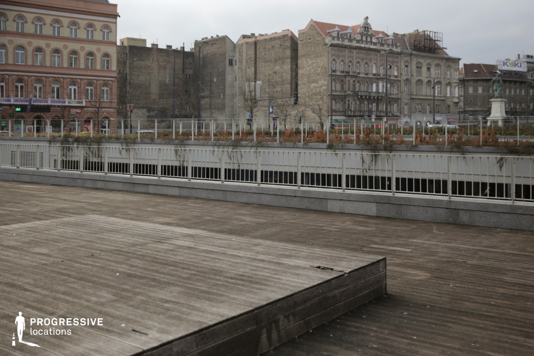 This image showcases an urban terrace featuring a broad wooden deck as its primary element. The deck’s weathered planks provide a textured, natural foreground that contrasts sharply with the historic and rugged background. A low railing topped with sparse vegetation encircles the terrace, softening its otherwise stark appearance.

In the background, the remnants of a historical cityscape tell a compelling story. On the left, a stately building with arched windows and ornate detailing conveys 19th-century elegance. However, the central portion reveals a striking void where a building once stood, now replaced by exposed firewalls and unfinished edges. These weathered walls and the gap between structures evoke a somber atmosphere, likely the result of war-time destruction. The stark, unadorned façades and the emptiness of the missing building create a sense of loss and resilience.

This setting is rich in visual and emotional contrasts. The juxtaposition of the terrace’s modern design and the haunting scars of historical upheaval offers a layered narrative. It is an ideal backdrop for projects seeking to highlight themes of time, history, and the interplay between destruction and renewal.