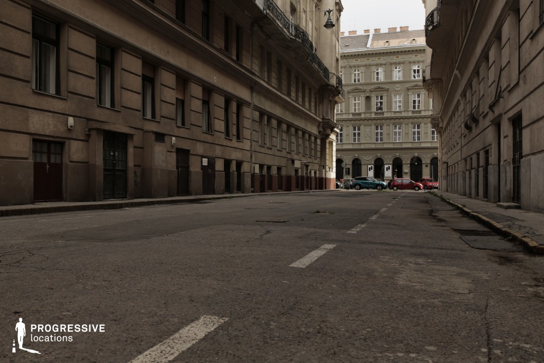 This image captures a quiet, narrow urban street framed by imposing historic tenement buildings. The façades on either side feature muted tones of beige and gray, with simple architectural details such as horizontal lines and recessed windows, creating a stark and somewhat austere appearance. The left side includes an iron-barred door and small entrances, reinforcing a sense of functionality and modesty in the design. The right side mirrors this simplicity, with minimal embellishments and uniformity. At the far end of the street, the view opens to a lighter, more ornate building with arched windows and decorative detailing, providing a contrasting focal point. The symmetry of the street and its gradual narrowing emphasizes a sense of confinement, while the visible cracks and imperfections on the asphalt contribute to an aged, weathered atmosphere. This setting, with its enclosed layout and subdued tones, evokes a feeling of isolation and timelessness. The lack of visible activity and the dominating architecture make it an excellent location for suspenseful or introspective scenes, particularly those exploring themes of urban decay or quiet tension.