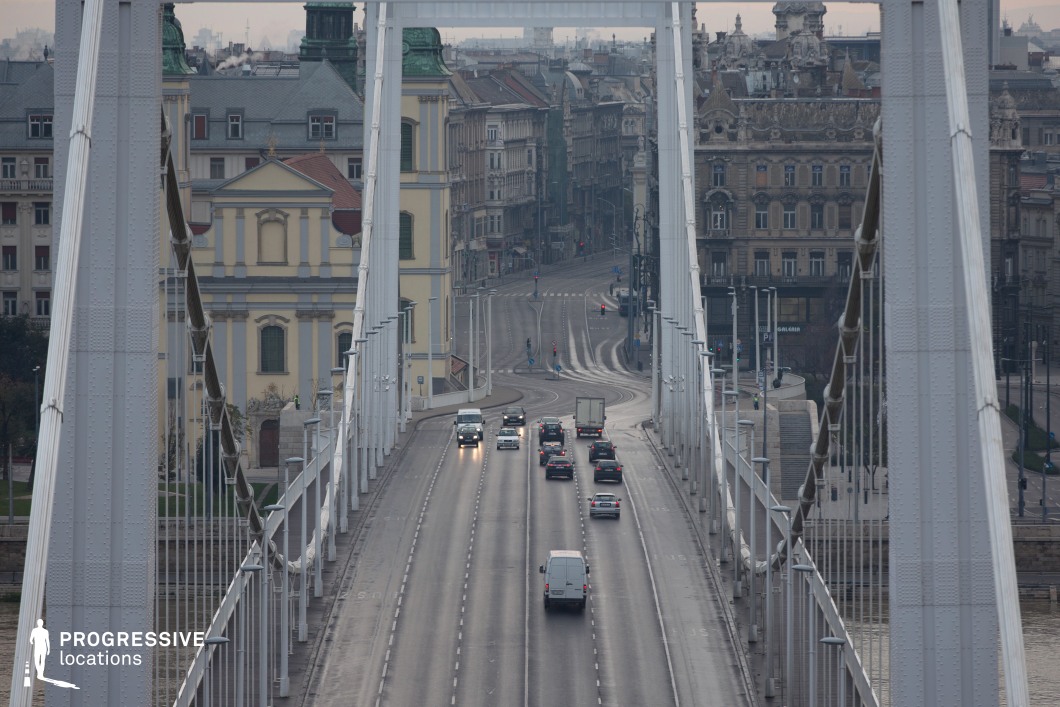 This striking image captures the Elisabeth Bridge in Budapest, elegantly framing a historic urban scene. The white suspension cables and modern structural elements of the bridge lead the eye towards the Baroque-style Inner City Parish Church in soft yellow, flanked by other richly detailed 19th-century buildings. The wide roadway of the bridge, carrying a steady flow of vehicles, underscores the dynamic connection between the modern and historical elements of the city. Beyond the bridge, the streets curve gracefully into the distance, lined with ornate facades that showcase a blend of Neo-Renaissance and eclectic styles. The juxtaposition of the clean lines of the bridge against the intricate details of the surrounding architecture highlights the harmony between old and new in this bustling urban landscape.