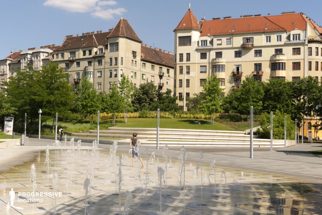 This image showcases a beautifully designed urban square that has been thoughtfully landscaped into a green park. The foreground features a lively modern fountain area with jets of water, perfect for capturing dynamic, playful scenes. Between the fountain and the backdrop of early 20th-century residential buildings with red-tiled roofs and stucco façades lies a lush green space, complete with manicured lawns, trees, and tiered seating. This greenery provides a relaxing atmosphere and serves as a visual bridge between the urban architecture and the interactive elements of the square. The combination of modern water features, park landscaping, and historic buildings makes this location ideal for productions focusing on urban outdoor activities, family scenes, or lifestyle narratives that highlight community spaces.
