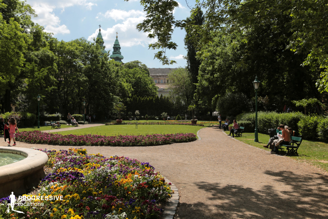 This image captures a beautifully maintained garden park that combines natural elements with subtle architectural details. The foreground is adorned with vibrant flowerbeds showcasing a variety of colors, bordered by neatly arranged stone curbs. A gently curving gravel pathway weaves through the park, connecting open lawns and providing an inviting route for leisurely walks. The park is surrounded by dense greenery, including tall trees and trimmed hedges, creating a serene and enclosed atmosphere. Elegant green iron street lamps are strategically placed along the paths, adding to the charm and blending seamlessly with the natural surroundings. In the background, the scene is crowned by two towering church spires, their cross-topped domes standing out against the leafy backdrop, evoking a sense of history and cultural significance. This setting’s harmonious layout and vibrant colors make it ideal for showcasing urban green spaces that balance relaxation and elegance. It offers a visually appealing and tranquil atmosphere, suitable for scenes that emphasize beauty, order, and the timeless appeal of well-kept gardens.