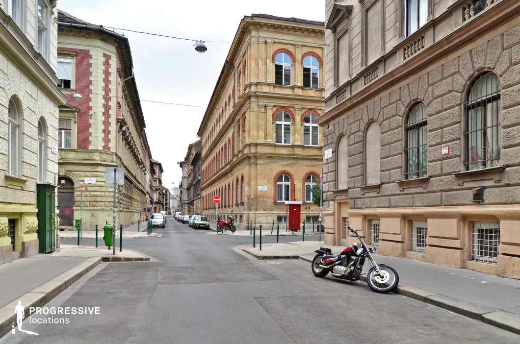 This serene city intersection features historic buildings with distinctive architectural elements, most notably the abundance of gracefully arched windows that give the facades a refined and timeless appearance. The corner building, with its striking combination of yellow and red brickwork, is further enriched by the symmetrical placement of these windows, adding rhythm and elegance to the design. The adjacent structures, with textured stone and subtle pastel tones, create a harmonious contrast while preserving a classic urban aesthetic. The clean streetscape and the attention to detail in the architecture make this location stand out as a unique and visually appealing urban setting, perfect for scenes requiring character and sophistication.