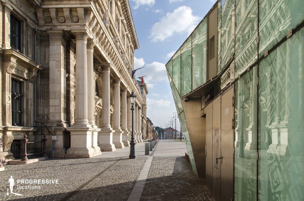 This image highlights the juxtaposition of classical and contemporary architecture. On the left, a historic building with grand columns, ornate stone carvings, and intricate details exudes timeless elegance. Its rhythm of repeating columns and symmetrical design reflects neoclassical grandeur, while the cobblestone pavement adds texture and depth to the foreground. On the right, a strikingly modern structure with reflective glass panels contrasts the historic façade. The glass reflects the classical architecture, blending the two styles visually and symbolically. The sharp, angular lines of the modern building and its clean metal surfaces emphasize functionality and contemporary aesthetics. This harmonious blend of old and new creates a visually engaging location, ideal for projects that explore themes of contrast, transition, or the merging of eras. The open perspective, with clear lines and ample space, provides versatility for cinematic or photographic compositions.