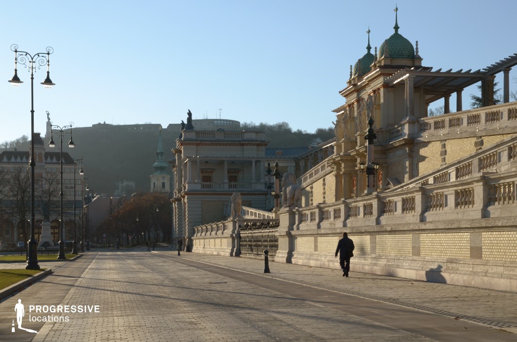 This image centers on a wide, paved street flanked by ornate historic architecture. The street’s cobblestone surface, bordered by neatly aligned grassy sections and classic wrought-iron street lamps, creates a formal and inviting atmosphere. The perspective draws the eye toward the distant background, where a baroque church spire and a hilltop monument add depth and cultural significance to the scene.

The symmetrical alignment of the street, with its expansive layout and unobstructed view, makes it a focal point for movement and interaction. While the right side features a stately terrace with detailed stonework and decorative elements, the street itself remains the dominant visual and functional element, offering ample space for dynamic activity or staging.

This location’s open composition and elegant surroundings make it ideal for scenes emphasizing grandeur, historical richness, or the flow of people and vehicles through a historic urban setting.