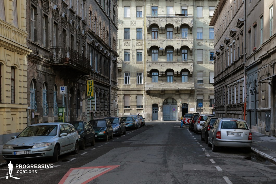 This image features a narrow urban street framed by historic buildings showcasing a mix of architectural styles. The central focus is on a prominent building in the background, characterized by symmetrical balconies, decorative reliefs, and a large arched doorway that draws the viewer’s attention. Its faded facade suggests a rich historical past, lending the location a sense of authenticity.

The buildings along the sides of the street exhibit worn yet intricate details, including wrought-iron balconies, arched windows, and textured stonework. The weathered appearance of these structures enhances the gritty and urban atmosphere, making the street feel lived-in and layered with history.

This enclosed urban setting, with its rich textures and architectural variety, is well-suited for scenes emphasizing tension, character development, or the interplay between elegance and decay. Its strong visual focus and depth make it versatile for cinematic and photographic compositions.