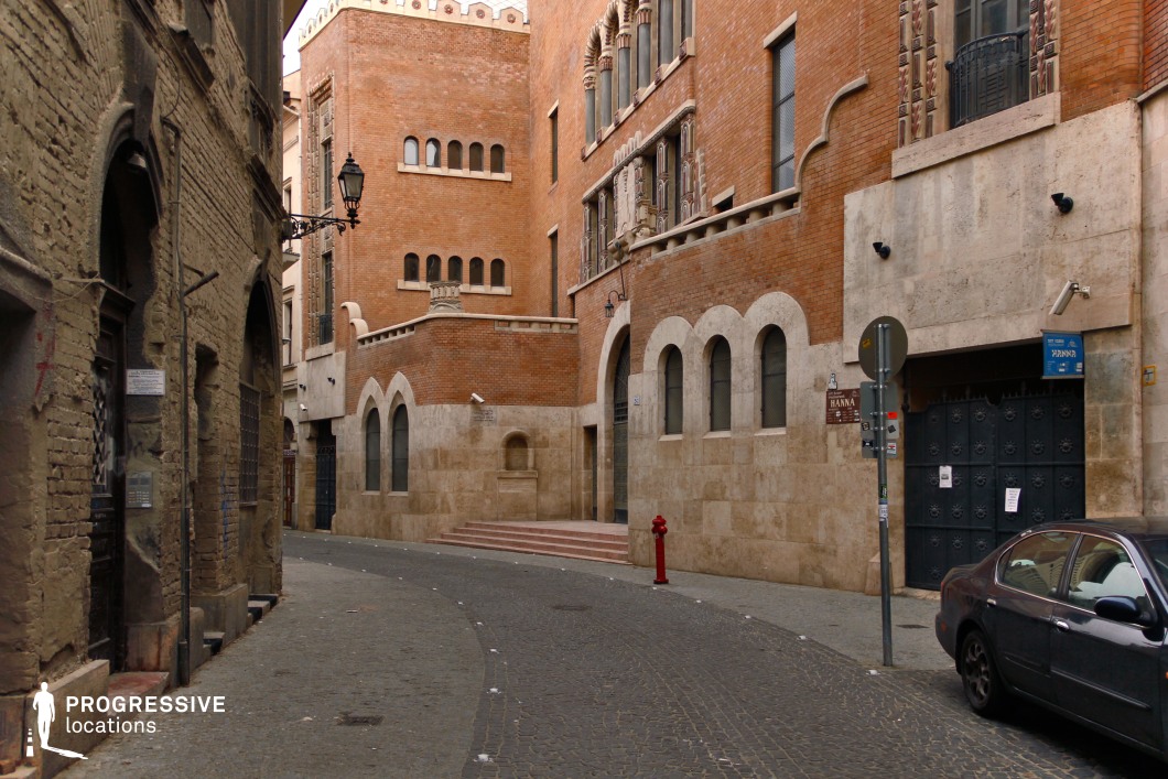 This intimate urban scene captures a winding cobblestone street nestled between historic buildings. The standout structure features richly colored brick facades with intricate architectural detailing, including arched windows and decorative stone elements that evoke a blend of Gothic and Moorish influences. The use of pale stone for the lower sections of the building and arched designs enhances its grandeur and visual appeal. On the opposite side, a more rugged and weathered brick structure creates a stark contrast, emphasizing the historical layers of the location. The narrow street, framed by ornate iron streetlamps and subtle embellishments, offers a secluded and timeless atmosphere. This setting is ideal for showcasing historical depth and architectural uniqueness in a compact, dramatic urban space.