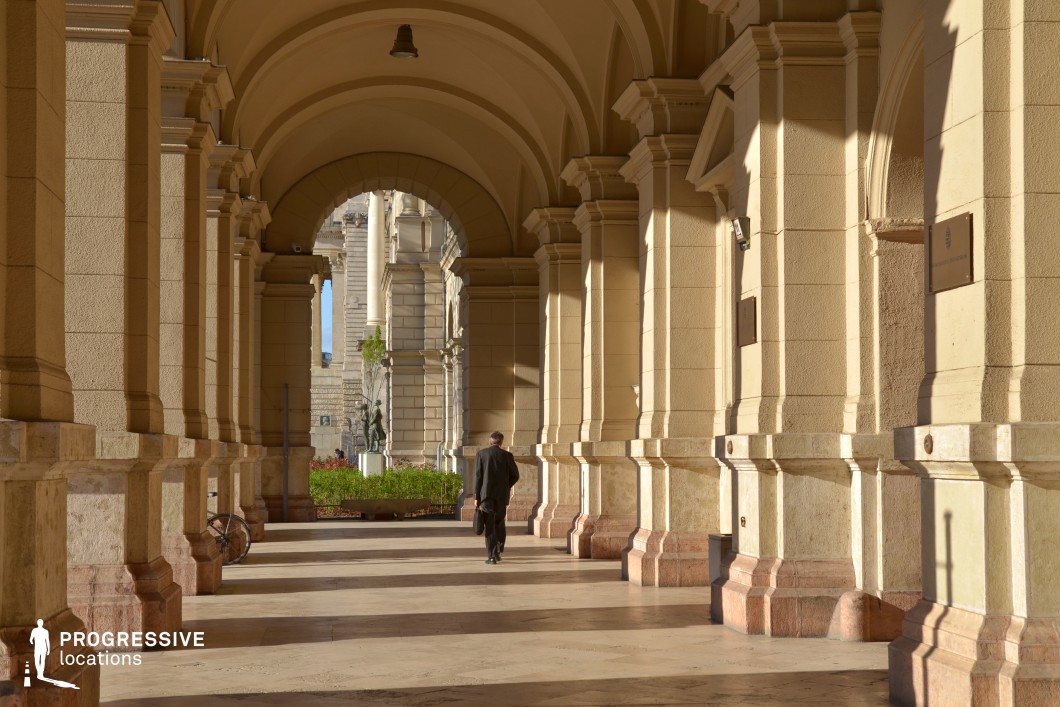 This image captures a grand arcade featuring an elegant series of classical columns and arches, designed with symmetry and precision. The light-colored stone structure reflects a warm, golden tone that enhances the sense of timeless sophistication. The arches frame views of a statue and manicured greenery at the far end, adding depth and visual interest to the space. The repeating pattern of columns creates a rhythmic play of light and shadow along the polished floor, emphasizing the arcade's architectural refinement. This location exudes an air of formality and grandeur, making it an ideal setting for elegant, cinematic, or historical scenes.