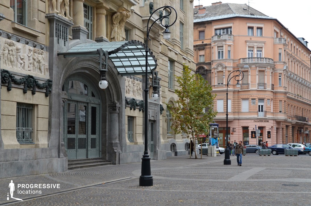 The focal point of this urban square is the grand Art Nouveau entrance, a masterpiece of architectural design. The arched doorway is framed by intricate ironwork, with detailed motifs that exemplify craftsmanship and elegance. Above the entrance, a glass and iron canopy extends outward, supported by sculptural elements that blend function with artistry. The facade is further adorned with stone reliefs depicting human figures and ornamental patterns, adding depth and historical character to the building. The entrance, with its imposing design and decorative details, is perfectly suited for scenes requiring a sophisticated or dramatic entryway. Surrounding the central building, the square is paved with decorative cobblestones, complemented by traditional street lamps and the warm tones of an adjacent peach-colored building, creating a balanced and inviting setting. This location is ideal for highlighting the grandeur of an iconic entrance in both film and photography.