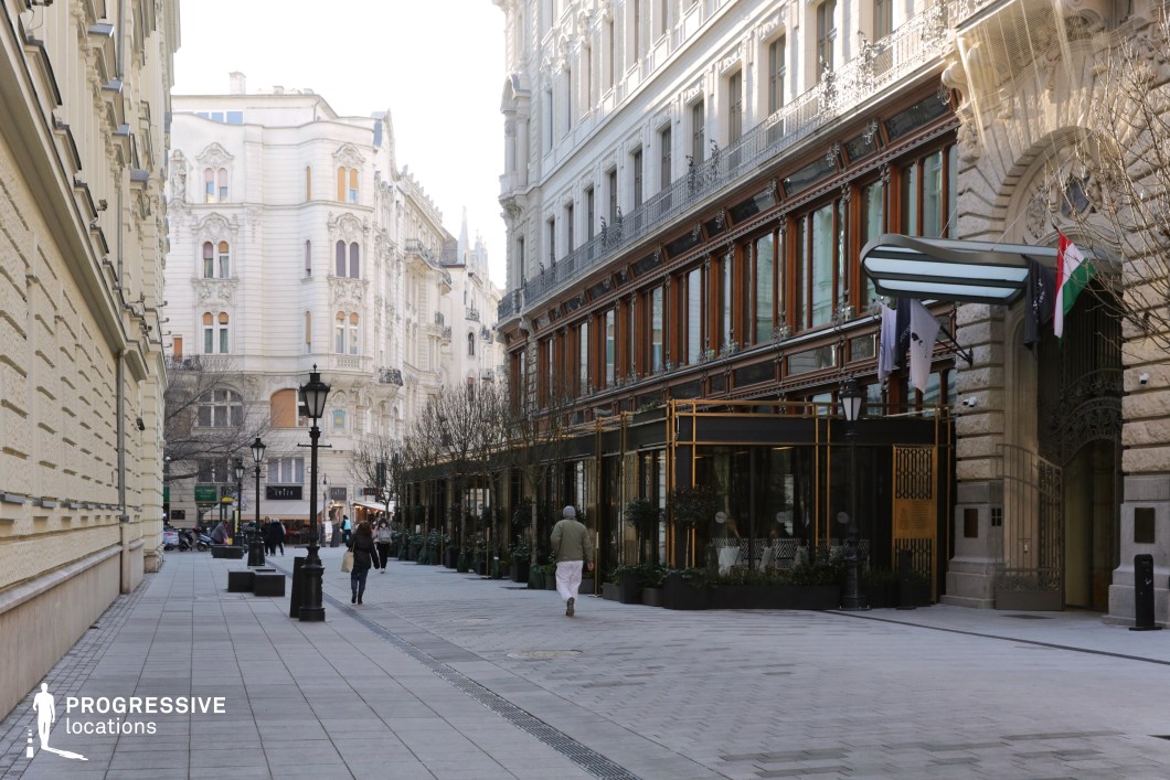 This location radiates sophistication with its seamless blend of classical architecture and modern design elements. The buildings' ornate facades with their intricate details frame the street, while the glass-enclosed café or restaurant stands out as a central feature. Unlike temporary signage, this structure is an integral, immovable part of the environment, serving as a dominant focal point.

The glass dining area, with its sleek metallic framework and large transparent panels, provides a striking contrast to the traditional stone facades and enhances the sense of exclusivity and elegance. Its presence adds a contemporary touch, reflecting the modern urban lifestyle within this historically rich setting.

The cast-iron candelabras with their dark finish and intricate detailing further enhance the classic feel of the street, providing continuity between the old and the new. Their robust, timeless design complements both the heritage architecture and the modern café structure.

This location is ideal for capturing scenes of refined city life, whether focusing on the vibrant activity of the café or the harmonious coexistence of historical and contemporary elements. The permanence of the café structure makes it a defining element of the scene, offering a sense of stability and uniqueness that sets it apart from more transient urban dining settings.